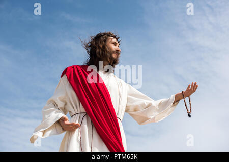 low angle view of Jesus in robe holding rosary and reaching hand against blue sky Stock Photo