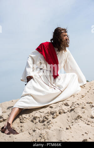 side view of Jesus in robe, red sash and crown of thorns holding rosary and squatting on sandy hill in desert Stock Photo