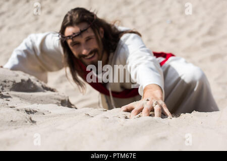 high angle view of smiling Jesus in robe, red sash and crown of thorns climbing sandy hill in desert Stock Photo