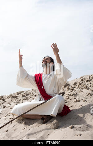 smiling Jesus in robe, red sash and crown of thorns sitting in lotus position with raised hands and talking with god on sand in desert Stock Photo