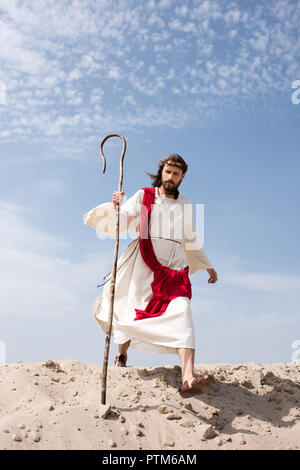 Jesus in robe, red sash and crown of thorns walking in desert with staff Stock Photo