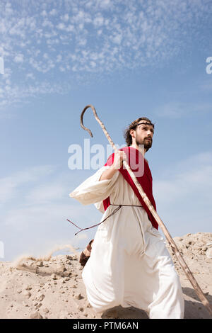 Jesus in robe, red sash and crown of thorns running in desert with staff Stock Photo