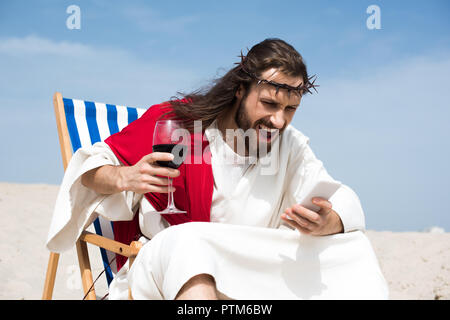 angry Jesus resting on sun lounger with glass of wine and screaming at smartphone in desert Stock Photo