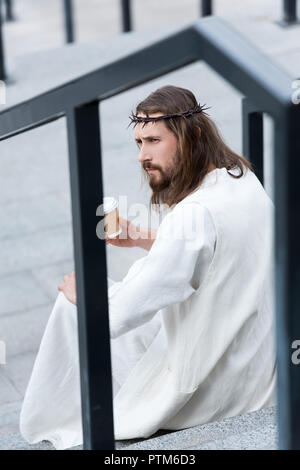 side view of serious Jesus in robe and crown of thorns sitting on stairs and holding disposable coffee cup on street Stock Photo