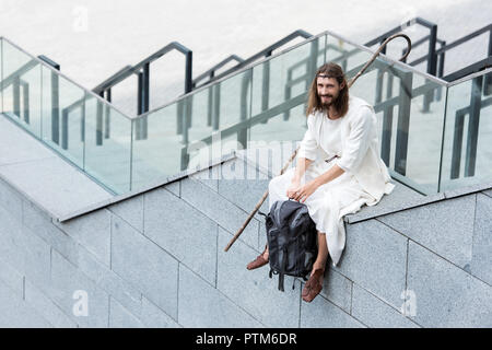 high angle view of smiling Jesus in robe and crown of thorns sitting on staircase side and holding bag Stock Photo