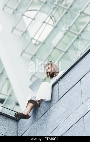 low angle view of Jesus in robe and crown of thorns using laptop while sitting on building wall Stock Photo