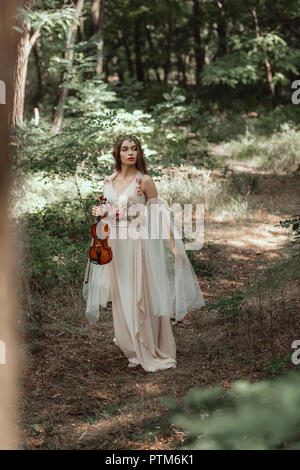 attractive mystic elf in elegant dress holding violin in beautiful forest Stock Photo