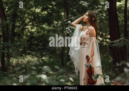 mystic elf character in flower dress holding violin in green forest Stock Photo