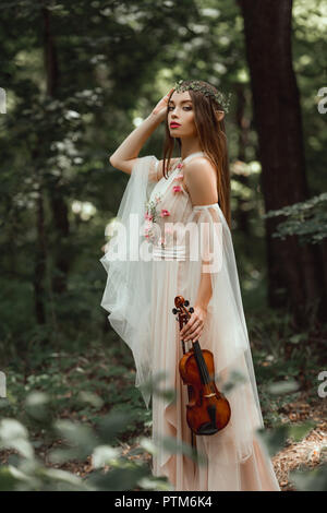 mystic elf in flower dress and floral wreath holding violin in forest Stock Photo
