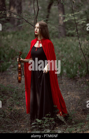 attractive mystic woman in red cloak holding violin in dark forest Stock Photo