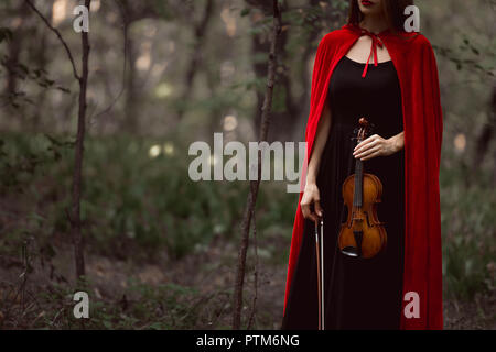 cropped view of elegant woman in red cloak holding violin in dark woods Stock Photo