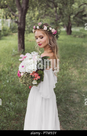 tender young bride in floral wreath and wedding dress holding bouquet of flowers outdoors Stock Photo
