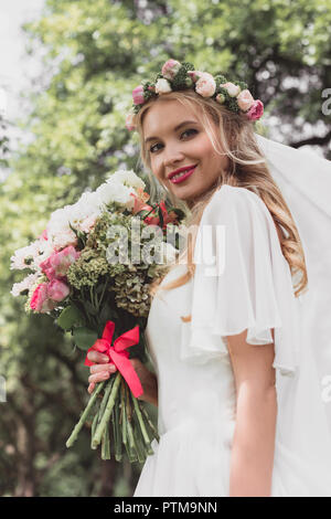 low angle view of beautiful young bride holding wedding bouquet and smiling at camera outdoors Stock Photo