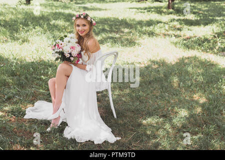 high angle view of happy young bride holding bouquet of flowers and smiling at camera while sitting on chair outdoors Stock Photo