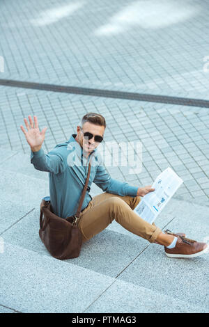smiling middle aged man sitting on stairs with newspaper and waving hand Stock Photo