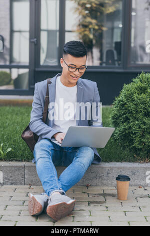 smiling asian teleworker using laptop while sitting on sidewalk with coffee to go Stock Photo
