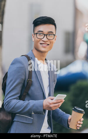 smiling asian man holding disposable cup of coffee and using smartphone Stock Photo