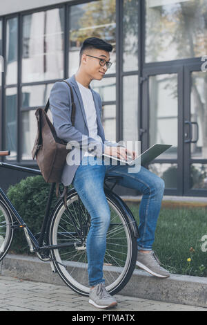 pensive asian teleworker with leather backpack using laptop while sitting on bicycle Stock Photo