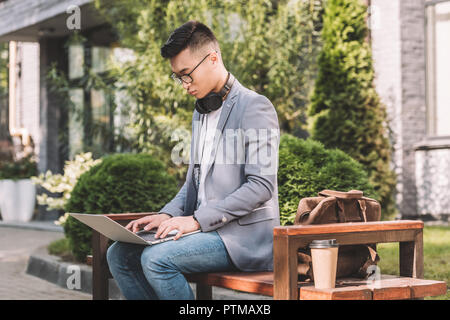 asian teleworker using laptop while sitting on bench with leather bag and coffee to go Stock Photo