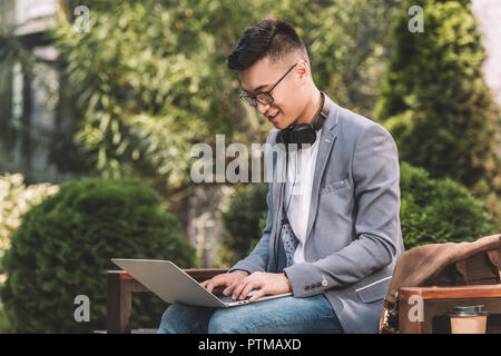 stylish asian freelancer in gray jacket working on laptop while sitting on bench Stock Photo