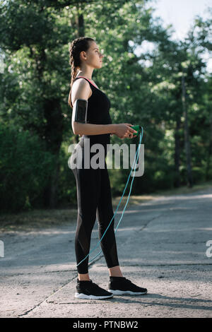 young sportswoman with smartphone in armband training with jump rope in park Stock Photo