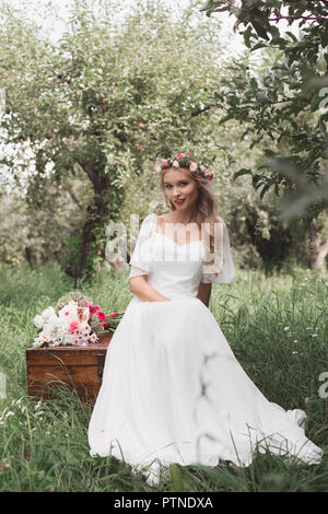 beautiful young bride sitting on vintage chest and looking at camera Stock Photo