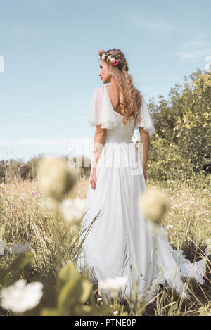 back view of beautiful young bride in wedding dress and floral wreath walking on field Stock Photo