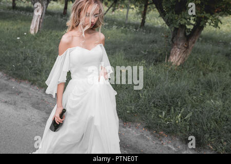 high angle view of upset young bride in wedding dress holding bottle of wine and walking in park Stock Photo