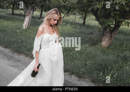 upset young bride in wedding dress holding bottle of wine and walking in park Stock Photo