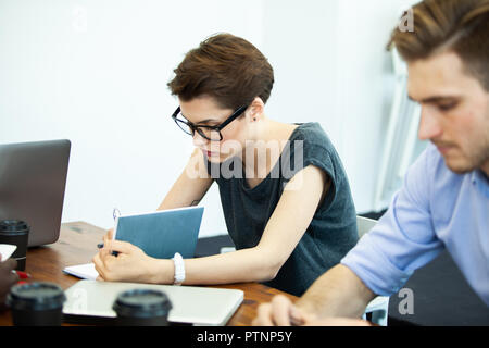 New solution every day. Confident young woman in smart casual wear working on laptop while sitting at her working place in office. Stock Photo