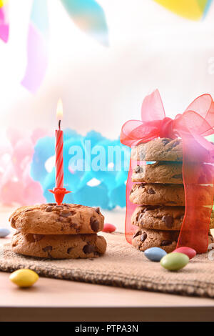 Birthday candle lit over chocolate cookies on a table with candy and colorful ribbons hanging in background. Concept of birthday. Front view. Vertical Stock Photo