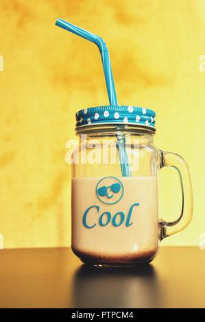 Cocoa milk drink in a big glass with handle, blue cover and straw on yellow background. In front is Cool and an emoticon written. Front view Stock Photo