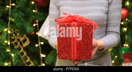 Female handing a red glitter square shape gift box wrapped with red ribbon bow to someone, blurred Christmas tree in background Stock Photo