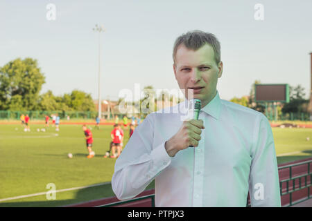 A male commentator in a white shirt with a microphone comments on football in the background of a football match Stock Photo