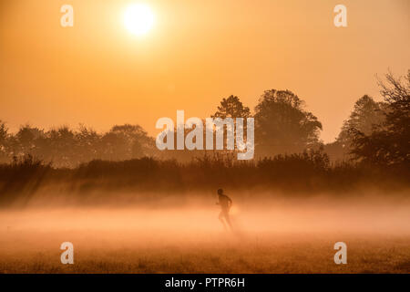 A man runs through the early morning mist as the sun rises over farmland near the village of Lower Wraxall in Wiltshire. Stock Photo