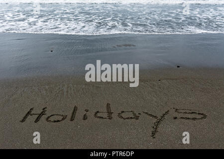 Waves lapping towards the word Holidays written in the sand. Stock Photo
