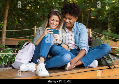 Excited young multiethnic couple talking while spending time together at the park, using mobile phone Stock Photo