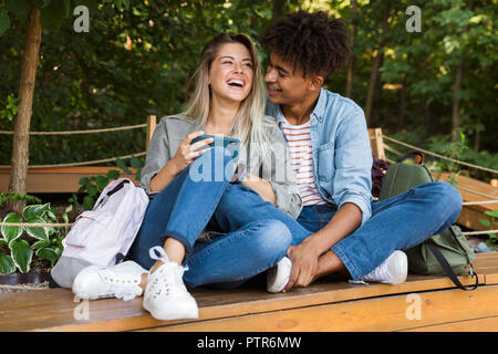 Laughing young multiethnic couple talking while spending time together at the park, using mobile phone Stock Photo
