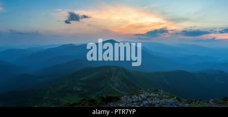 Fantastic sunrise or sunset over green mountain ridge covered with dense blue fog. Bright orange sun raising in  soft cloudy sky over distant horizon. Stock Photo