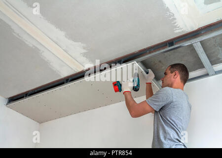 Young man in usual clothing and work gloves fixing drywall suspended ceiling to metal frame using electrical screwdriver on ceiling insulated with shi Stock Photo