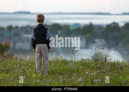 Back silhouette of young blond child boy standing alone on grassy hill top on spring or summer day on distant foggy blurred blue view of village house Stock Photo