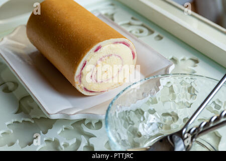 Arctic roll cake on a plate on a tray next to a glass bowl with spoons and fork in it. Dessert. Stock Photo