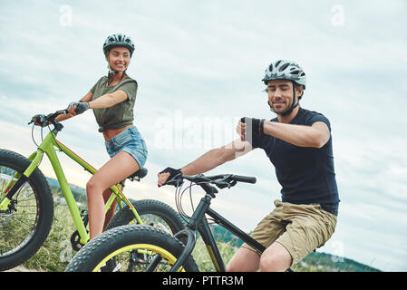 Young couple are cycling on fatbikes at green hills. Man looks at his wristwatch. Side view Stock Photo