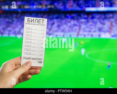 A woman's hand holds a bookmaker's ticket against the backdrop of a game of football, sports betting, the bookmaker's office. Билет букмекерский офис -Ticket bookmakers office, ставка-bet, коэффициент-ratio, общий выигрыш - total win Stock Photo