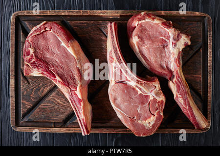 three Dry aged raw tomahawk beef steaks on an old rude wooden cutting board, close-up, flat lay, view from above Stock Photo