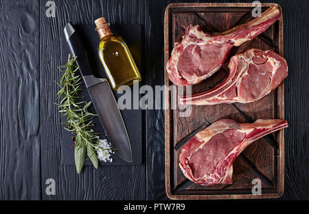 three Dry aged raw tomahawk or cowboy beef steaks on an old rude wooden cutting board on a black table with knife, spice and herbs, close-up, flat lay Stock Photo