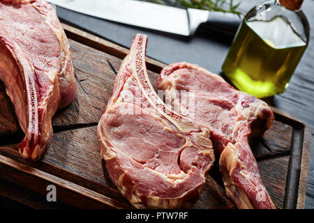 three Dry aged raw tomahawk or cowboy beef steaks on an old rude wooden cutting board on a black table with knife, spice and herbs, close-up, Stock Photo