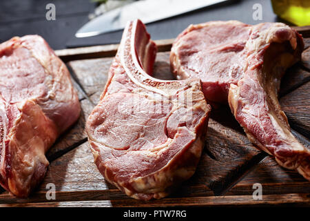 three Dry aged raw tomahawk or cowboy beef steaks on an old rude wooden cutting board on a black table with knife, spice and herbs, close-up, view fro Stock Photo