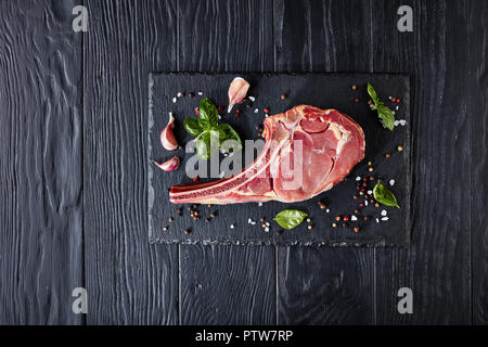 Dry aged raw tomahawk or cowboy beef steak on a stone board  with spices, peppercorns, garlic cloves and basil, close-up, flat lay, view from above, Stock Photo