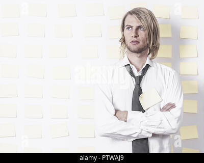 Junger Mann steht in Hemd und Krawatte vor Postit-Wand, verschraenkte Arme (model-released)